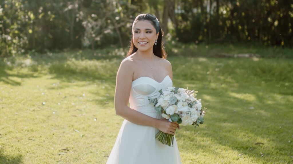 Noiva radiante com maquiagem profissional duradoura, penteado elegante e buquê de flores brancas em casamento ao ar livre, resultado do trabalho de visagismo da Mariana Botelho no Rio de Janeiro