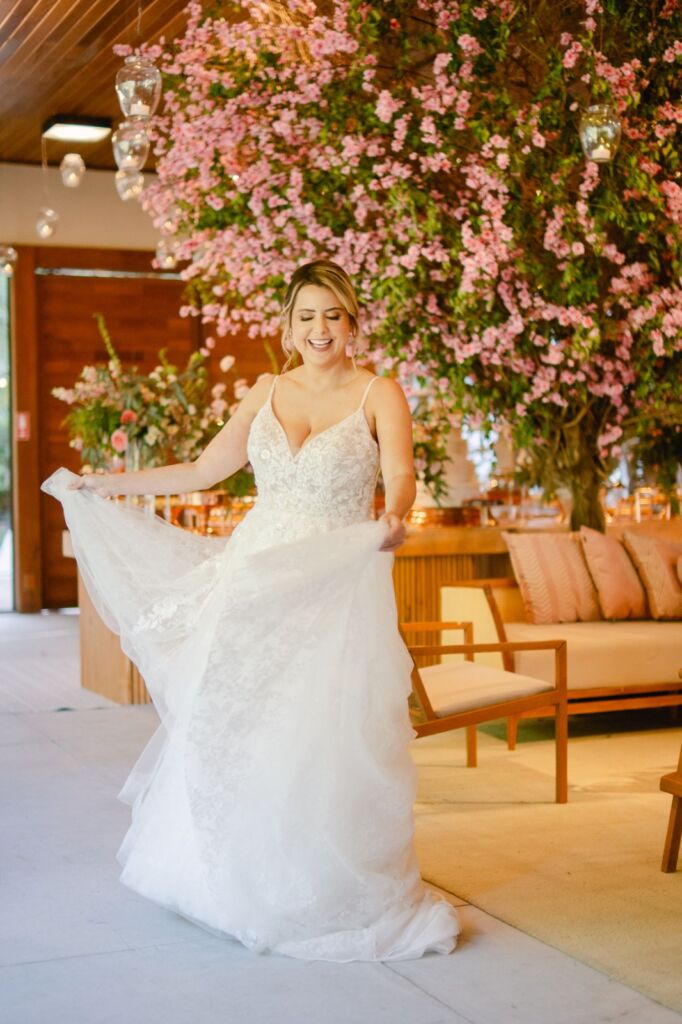 Noiva radiante sorrindo em movimento com vestido fluido de renda sob decoração exuberante de flores rosas suspensas, maquiagem duradoura visagismo Mariana Botelho