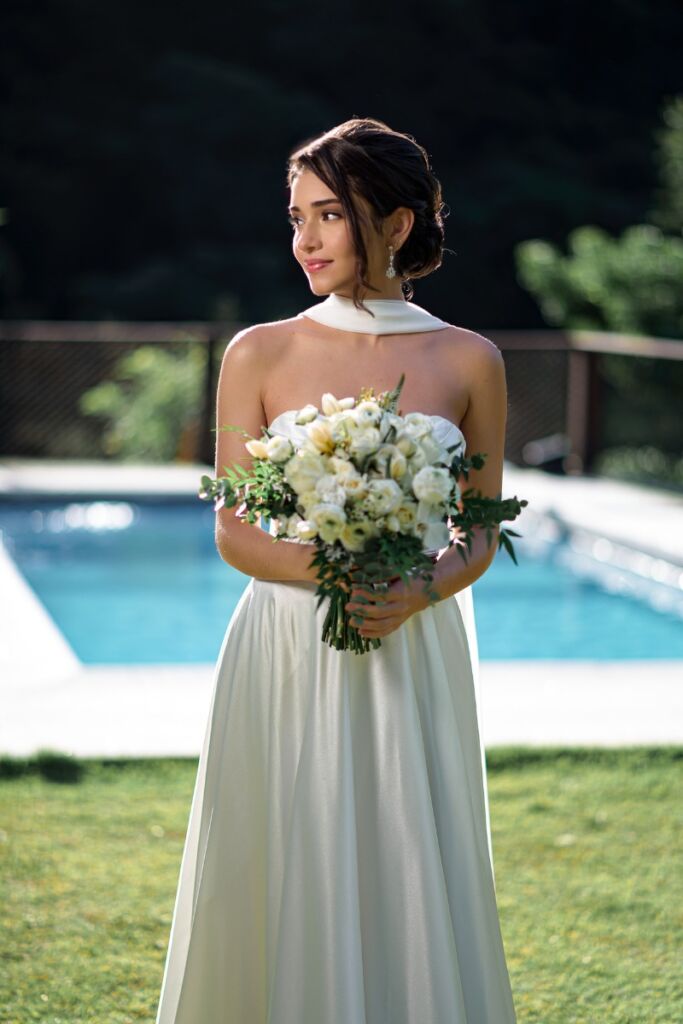 Noiva jovem sorrindo suavemente, segurando um buquê de flores brancas e folhagens, com maquiagem iluminada e penteado preso, em cenário externo com luz solar